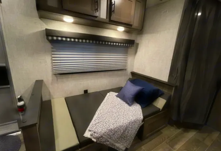 RV rental interior features a convertible gray dinette bed with pillows, blanket, window with blinds, and overhead cabinets.. Grandriver 25BH 2021