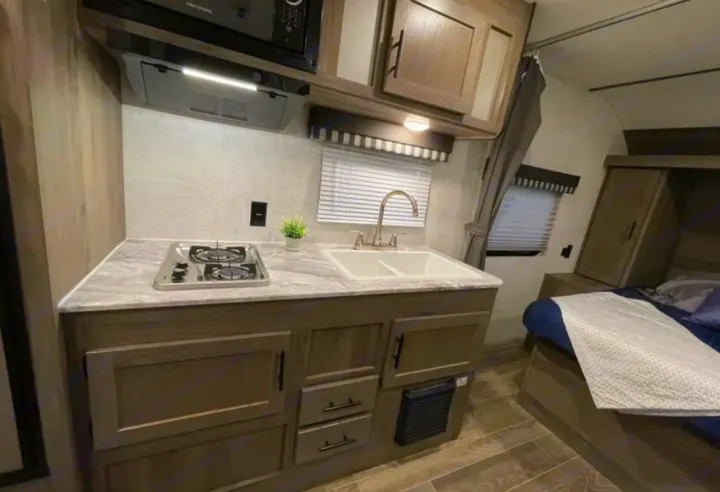 Inside an RV rental: a modern, compact kitchen with sink, stove, and microwave, leading to a cozy bedroom area.. Grandriver 25BH 2021