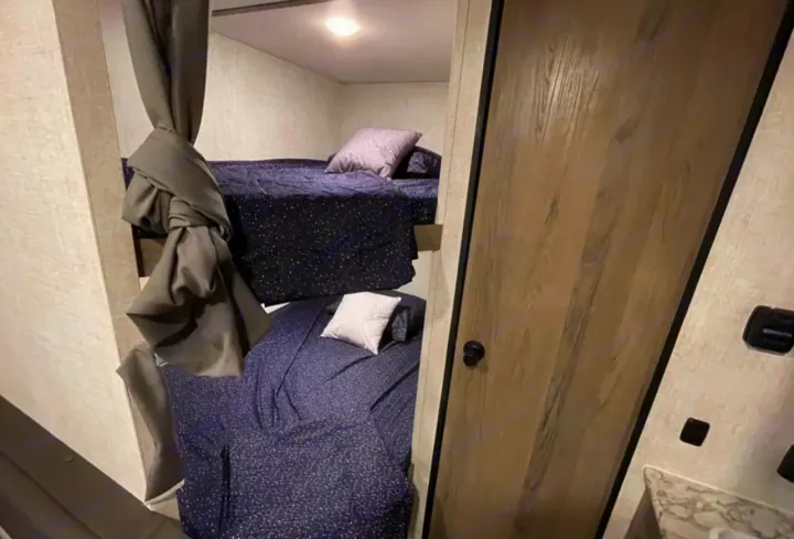 Inside an RV rental, showing a compact bunk bed setup with dark blue bedding, privacy curtain, and adjacent door.. Grandriver 25BH 2021