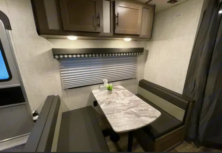 Cozy RV rental dinette interior: grey marble-look table, dark benches, overhead cabinets, & window blinds.. Grandriver 25BH 2021