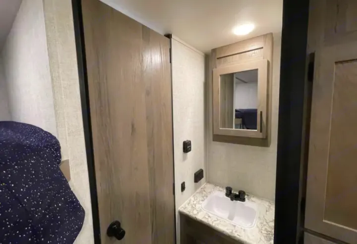 RV rental interior: A compact bathroom with a white sink and mirrored cabinet, next to a bed with a dark blue blanket.. Grandriver 25BH 2021