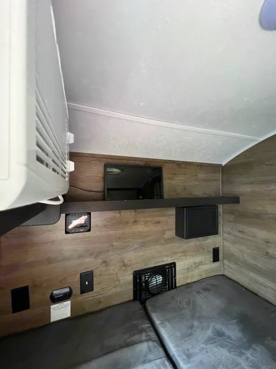 View of an RV rental interior with wood-grain walls, showcasing an overhead AC unit, wall-mounted TV, control panel, and beds.. Braxton Creek Bushwhacker 2021
