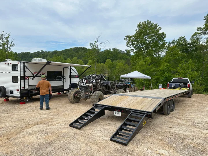 Camping with an RV rental, a large flatbed trailer, and various off-road vehicles parked on a dirt lot amidst green hills.. Forest River Wildwood FSX 2021
