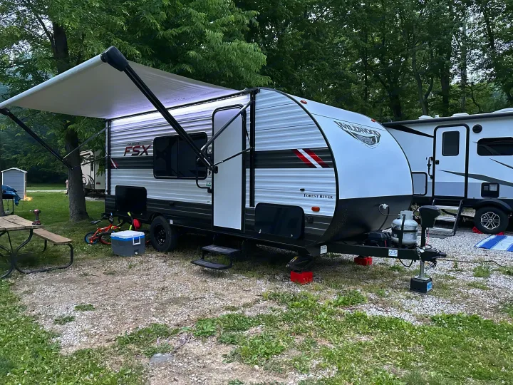 A white Wildwood FSX travel trailer, likely an RV rental, set up at a gravel campsite with its lighted awning extended.. Forest River Wildwood FSX 2021