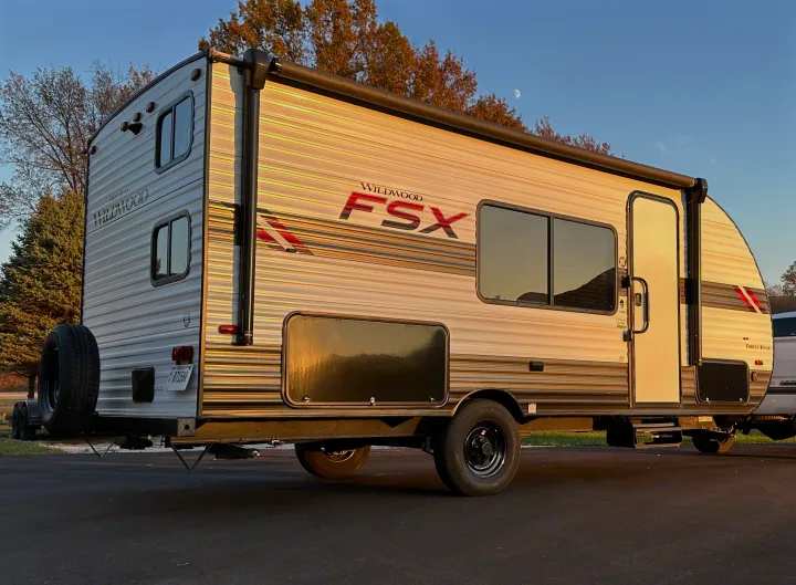 Wildwood FSX travel trailer, an ideal rv rental, is parked outdoors. The side shows windows & a spare tire, under a blue sky with a moon.. Forest River Wildwood FSX 2021