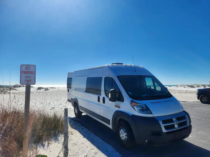 White RV rental van parked on a sunny beach, next to a "No Parking Any Time" sign. Ocean visible in the distance.. Dodge 1500 2014