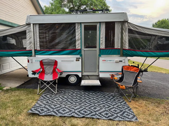 This pop-up RV rental is set up outdoors with camping chairs & a patterned mat, ready for a relaxed camping experience.. Coleman Americana Utah 1995