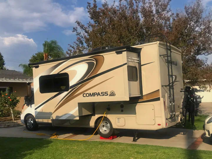 Beige RV rental, a Compass AWD model, parked on a driveway with an extended slide-out, bike rack, and power hookup.. Thor Compass 2021