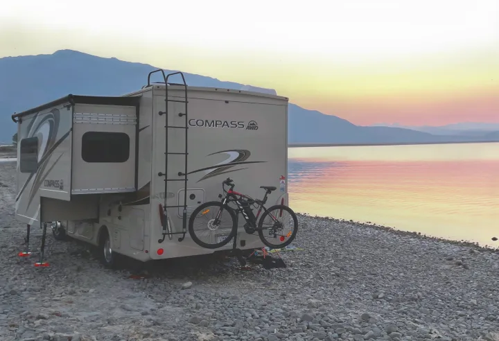 An RV rental, complete with bike, parked lakeside at sunset, showcasing serene nature and travel possibilities.. Thor Compass 2021