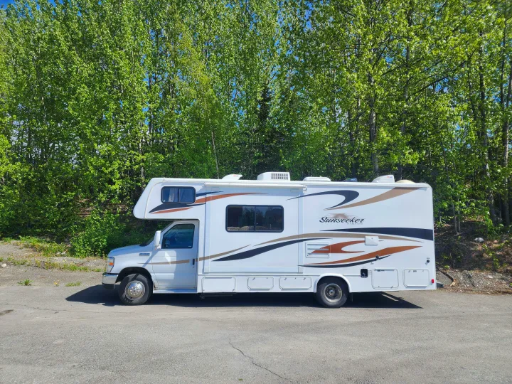 White Sunseeker RV rental (motorhome) parked on asphalt with vibrant green trees in the background.. Forest River Sunseeker 2012