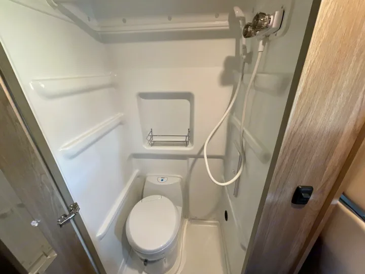 Compact RV rental wet bath with white toilet, mounted showerhead, and small shelving, demonstrating space-saving design.. Winnebago Solis 2021