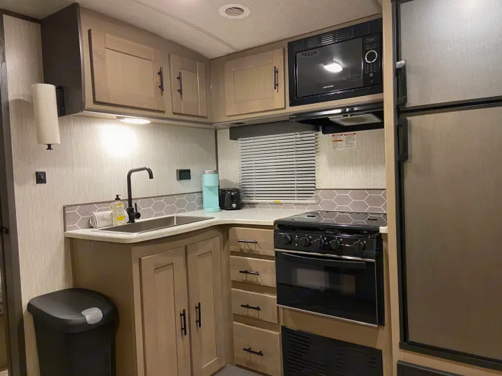 Modern RV rental kitchen featuring a sink, stove, microwave, fridge, light wood cabinets, and a hexagon tile backsplash.. Shadow Trailers 277BHS 2020