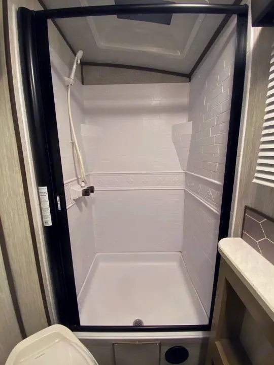 A well-equipped, spotless shower inside an RV rental. Features white faux-tiled walls, handheld showerhead, and modern controls.. Shadow Trailers 277BHS 2020