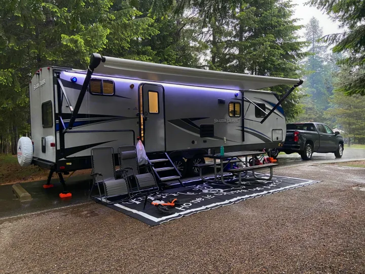 An RV rental parked at a wet campsite with a truck, extended awning, and LED lights, ready for forest camping.. Shadow Trailers 277BHS 2020