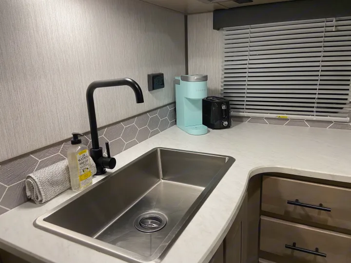 A sleek rv rental kitchen with a stainless sink, black faucet, light blue Keurig, and toaster. Gray and white accents.. Shadow Trailers 277BHS 2020