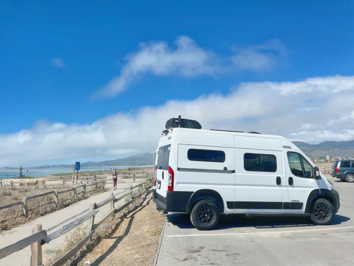 White rv rental camper van parked by a scenic coastal path under a blue sky, with a person walking. Ideal for outdoor travel.. Winnebago Solis Pocket 2022
