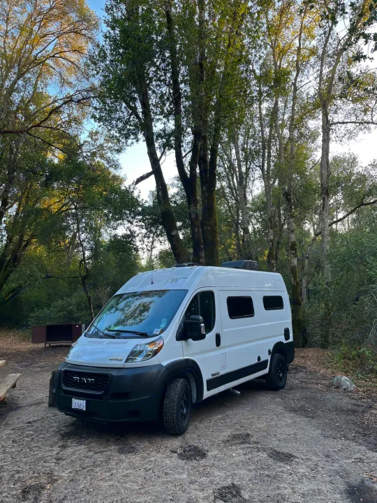 White RV rental parked at a serene wooded campsite, surrounded by tall trees and nature.. Winnebago Solis Pocket 2022