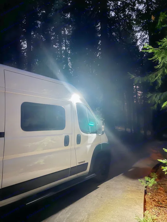 White RV rental van parked at night, illuminated by bright side lights against a dark forest background.. Winnebago Solis Pocket 2022