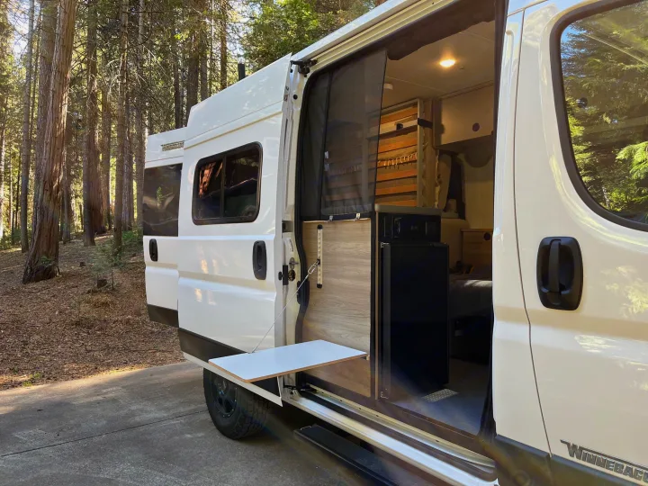 Discover freedom with this RV rental. White camper van in a sunny forest, its side door open to reveal a cozy interior and pull-out table.. Winnebago Solis Pocket 2022