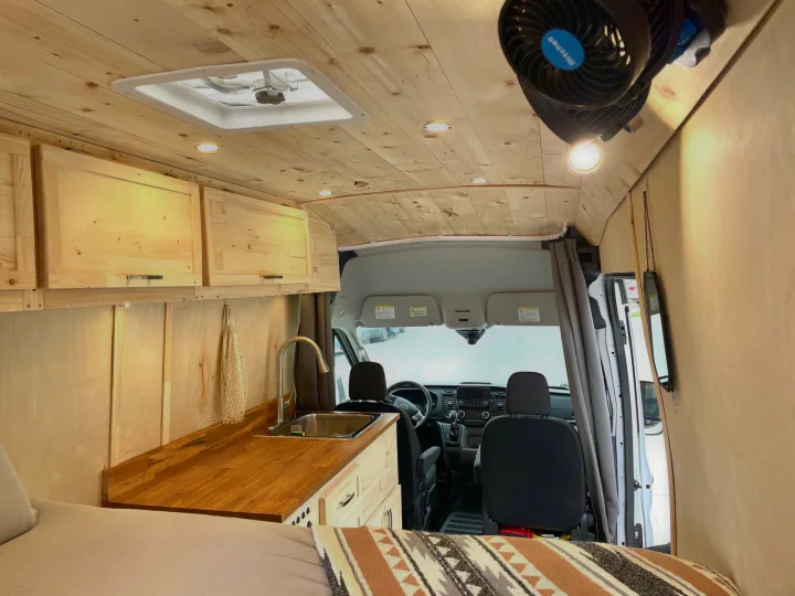 This RV rental van boasts a cozy wooden interior with a kitchen, sink, overhead fan, comfy bed, and front cabin. Ideal for travel.. Ford Transit Custom 2022
