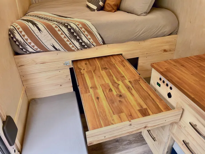 RV rental interior features a cozy bed with a patterned blanket, a pull-out wooden table, and light wood walls.. Ford Transit Custom 2022