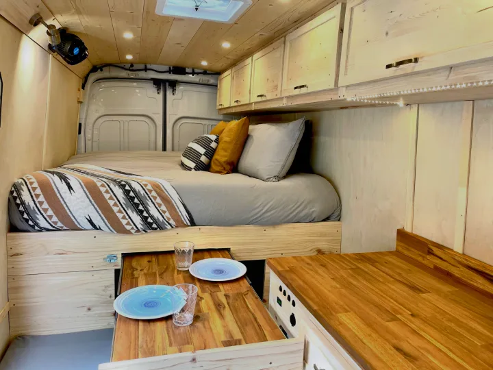 This RV rental showcases a custom wood interior with a comfy bed, pull-out table, and built-in storage.. Ford Transit Custom 2022