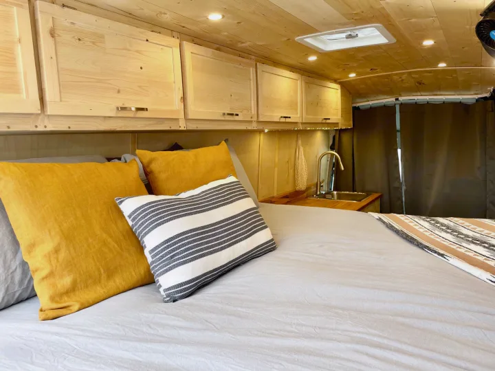 Comfortable RV rental interior with a bed, yellow & striped pillows, wooden cabinets, and a compact kitchen sink.. Ford Transit Custom 2022