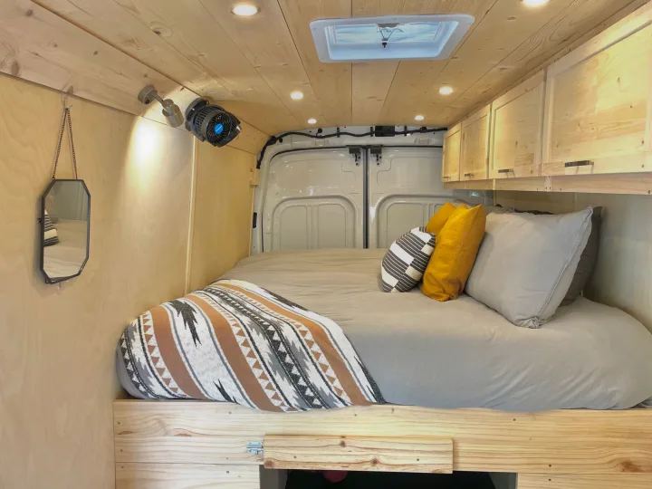 RV rental camper van interior: a cozy bed with patterned blanket, wooden walls/ceiling, overhead cabinets, and warm lighting.. Ford Transit Custom 2022