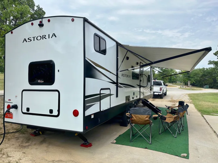 Astoria travel trailer RV rental parked at a campsite, with an extended awning and chairs on a green mat.. Dutchmen Astoria 2023