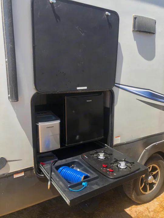 RV rental outdoor kitchen: open hatch shows fridge, ice maker, pull-out stove, and sink. Perfect for camping meals.. Forest River Cherokee Arctic Wolf 2020