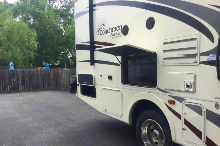 Side view of a cream-colored Coachmen Pursuit RV rental, showing open storage and an outdoor setting.. Coachmen Pursuit 27DS 2019