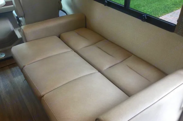 An RV rental interior showing a light beige convertible sofa that unfolds into a bed, with a window above.. Coachmen Pursuit 27DS 2019
