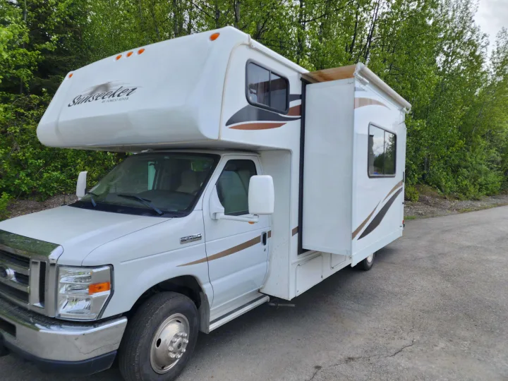 White Sunseeker RV rental parked on asphalt, with its slide-out extended, surrounded by green trees.. Forest River Sunseeker 2012