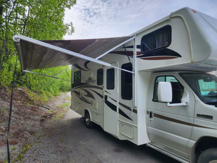 Experience nature with this spacious RV rental, parked on a gravel path with its awning extended under green trees.. Forest River Sunseeker 2012