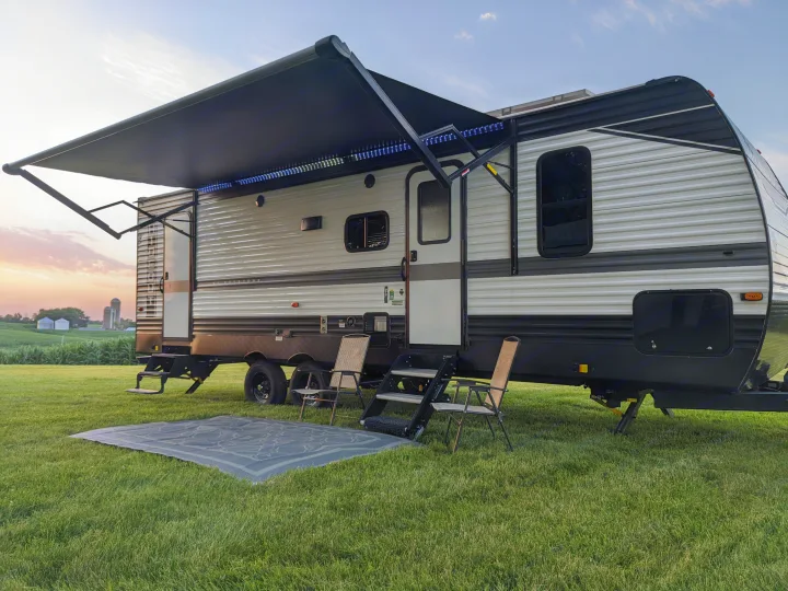 RV rental at sunset. A travel trailer with an extended, lit awning, outdoor chairs, and a mat on green grass.. Keystone RV Hideout 2022