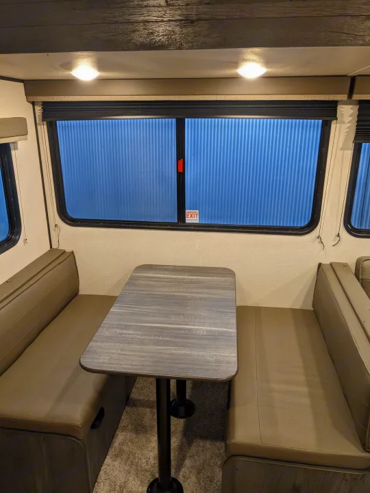RV rental interior featuring a dinette with a grey wood-look table, comfy seating, and windows, one with an "Emergency Exit" sign.. Keystone RV Hideout 2022