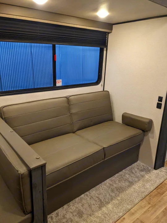 RV rental interior: a grey sofa, window with emergency exit sign, and carpeted floor under bright lights.. Keystone RV Hideout 2022