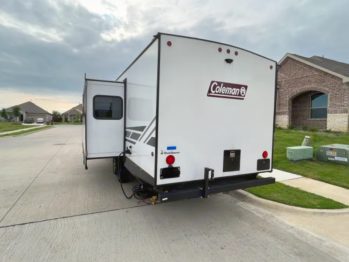 Rear view of a white Coleman RV rental with its slide-out extended, parked on a residential street under a cloudy sky.. Dutchmen Coleman Light 2022