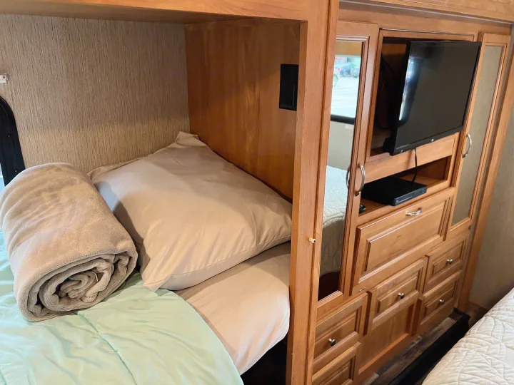 RV rental bedroom: A cozy bunk bed with light sheets and blanket, beside an entertainment center with a TV and media player.. Nexus Phantom 32P 2019
