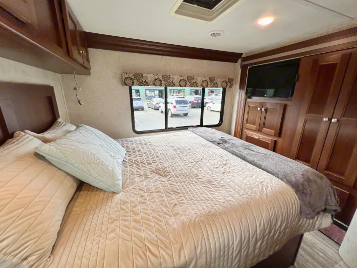 Cozy RV rental bedroom featuring a comfortable bed, overhead cabinets, TV, and a window view of a parking lot.. Forest River Forester 2019