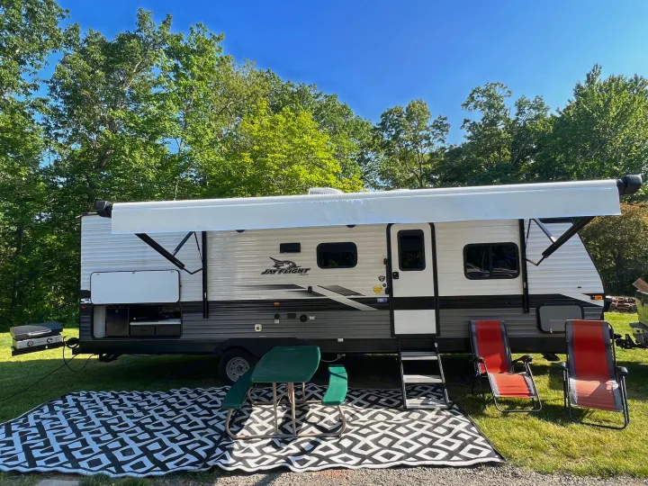 RV rental travel trailer with an extended awning, outdoor kitchen, black/white mat, picnic table, and chairs at a lush campsite.. Jayco Jay Flight SLX 2022
