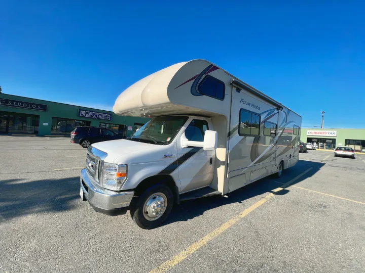 Sunny day shot of a white Four Winds Class C RV rental parked in a lot near businesses.. Thor Motor Coach Four Winds 2019