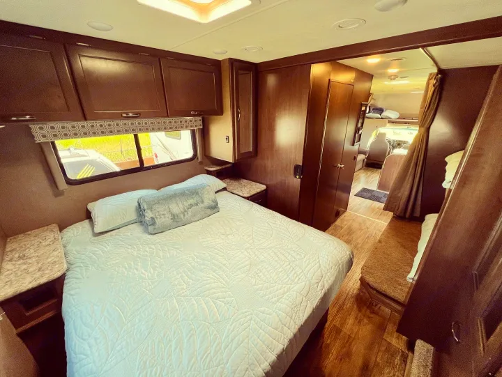 Inside an **RV rental**: cozy bedroom with light bedding, overhead storage, and a hallway revealing bunk beds and the front cab.. Thor Motor Coach Four Winds 2019