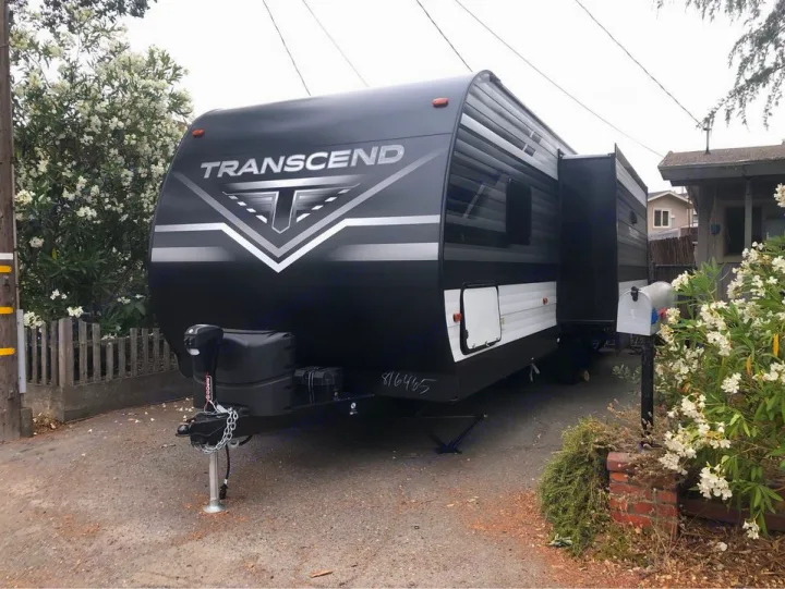 Modern black Transcend RV rental travel trailer parked in a driveway. Features a slide-out; surrounded by white blooming bushes.. Grand Design transcend 2021