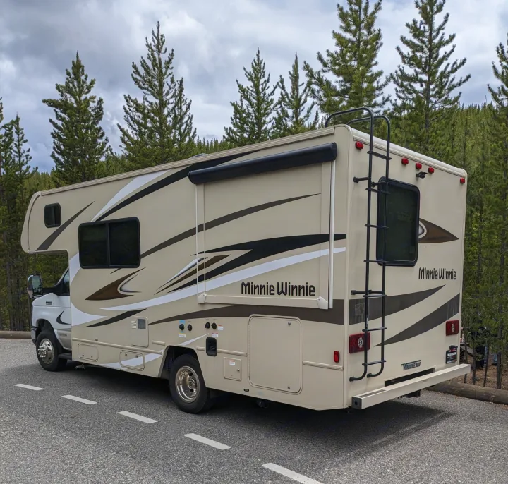 A beige Minnie Winnie RV rental is parked on asphalt with pine trees and a cloudy sky in the background, ready for an adventure.. Winnebago Minnie Winnie 2022