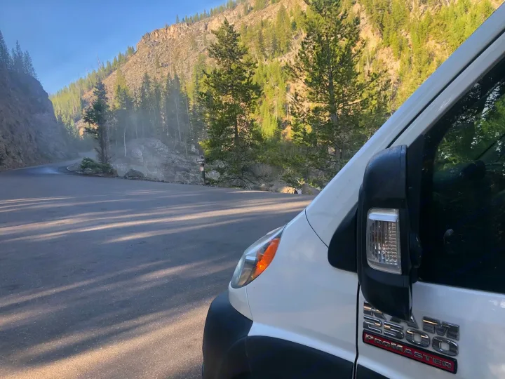 White RV rental van on a mountain road overlooking misty cliffs and pines near Firehole Falls. Scenic nature trip begins!. Dodge RAM Promaster 2500 2019