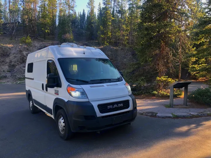 White Ram ProMaster camper van, an RV rental, parked on a paved lot amidst a pine forest and rocky hill.. Dodge RAM Promaster 2500 2019
