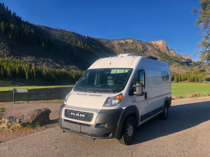 White RAM ProMaster rv rental van by a river, with lush green fields and forested mountains under a clear blue sky. Ideal for nature travel.. Dodge RAM Promaster 2500 2019