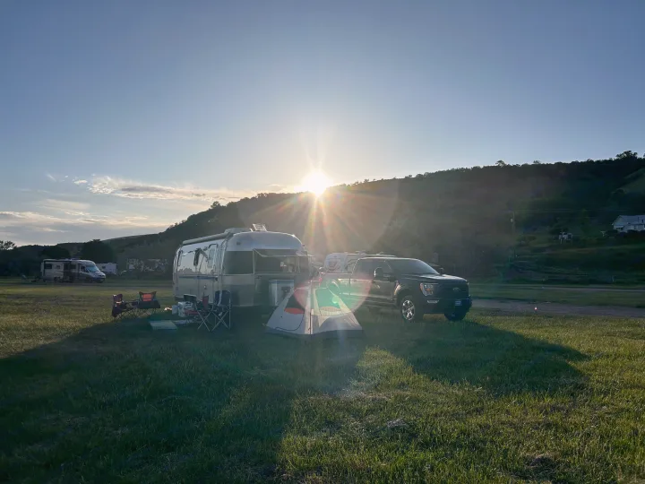 Sunset view of an Airstream RV rental, truck, and tent at a grassy campsite with hills.. Airstream Excella 2000