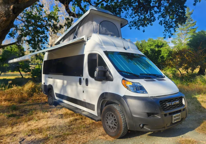 White pop-top RV rental with awning extended, parked in nature. Ready for your next camping adventure!. Thor Tellaro 2023